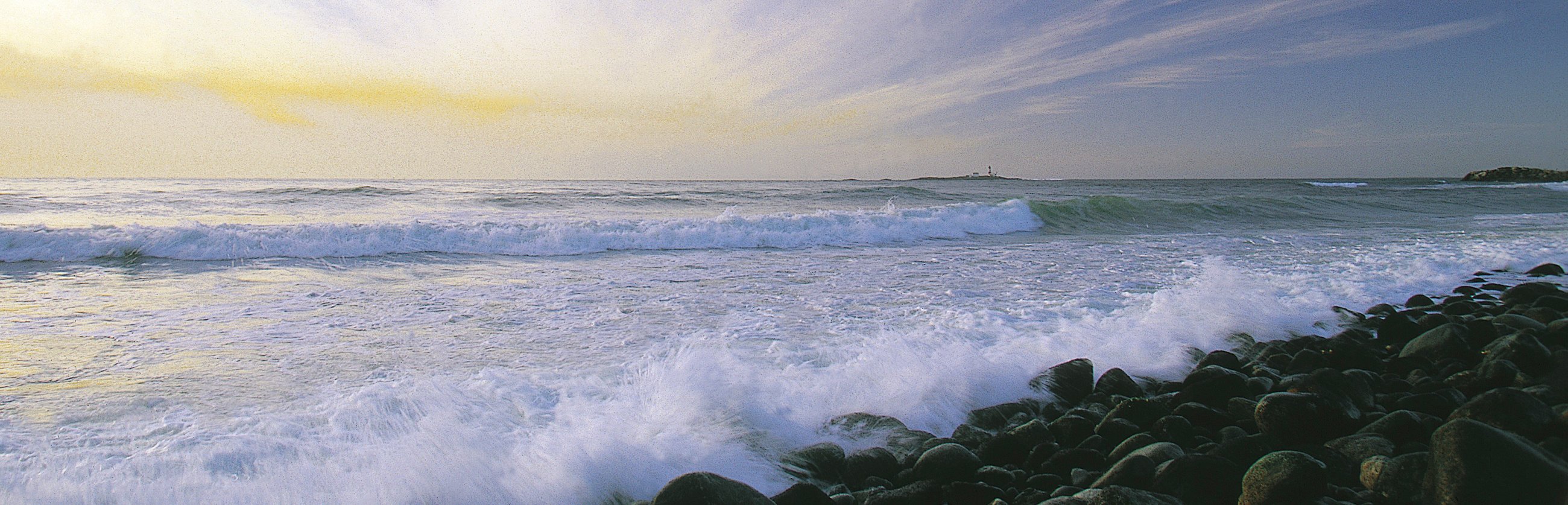 Bølger slår mot grov rullesteinstrand. Himmelen er gul av solnedgang og det er et lite fyr i horisonten